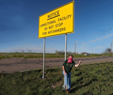 Masked Hitchhiker Next To A Road Sign that Says : 